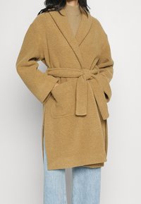 Accappatoio marrone in pile testurizzato con collo a scialle, vita con cintura e tasche laterali, abbinato a jeans azzurri.