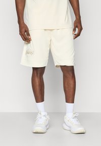 Cremefarvede træningsshorts med opslåede kanter, der har et Los Angeles Lakers-logo i tan. Bæres med hvide sneakers og sokker.