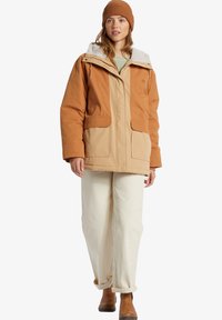 Brun och orange isolerad jacka med en foderhuva i päls, två framfickor, kombinerad med ljusbeiga byxor och bruna stövlar.