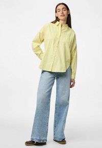 Camicia button-up a righe gialle con tasca, abbinata a jeans a gamba larga azzurri e scarpe sportive marroni. Tessuto morbido con vestibilità rilassata.
