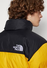 Sort og gul pufferjakke med høj krave, der har et hvidt "The North Face" logo på venstre skulder. Textureret stof.