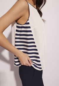 Top bez rękawów z jednolitym kremowym materiałem z jednej strony i niebiesko-białymi paskami z drugiej, w luźnym kroju i zaokrąglonym hemem.