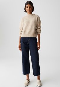 Donna in piedi, con un maglione a coste color crema, jeans cropped larghi blu scuro e scarpe slip-on bianche, su uno sfondo grigio neutro.