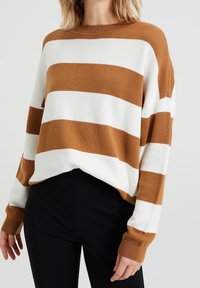Brun och vit randig tröja med lös passform, långa ärmar, rund halsringning och ribbade muddar. Buren över svarta byxor.