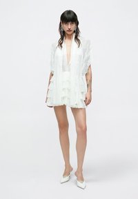 Biele vrstvené minišaty z priehľadnej čipkovanej látky, s volánmi, hlbokým výstrihom do V a širokými rukávmi. Štýlovo doplnené bielymi špicatými topánkami.