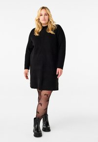 Svart kabelstickad klänning med rund halsringning, långa ärmar och avslappnad passform; tillsammans med genomskinliga svarta tights med rosettmönster och chunky svarta stövlar.