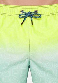 Pantaloni scurți de înot verzi și galbeni cu un design în gradient, având o talie cu șnur și un model repetitiv de text alb. Textură netedă.