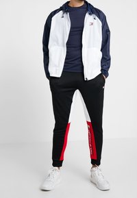 Homme portant une veste bleu marine et blanche, une chemise bleu marine, un pantalon noir avec des bandes blanches et rouges, et des baskets blanches, se tenant les mains dans les poches.