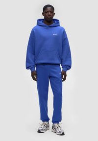 Felpa blu con tasca a marsupio e logo, abbinata a pantaloni jogger blu coordinati. Materiale morbido in cotone misto, texture soffice e polsini elasticizzati.