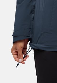 Veste imperméable marine avec ourlet ajustable à cordon de serrage, tissu lisse, manches longues et fermeture sécurisée aux poignets.