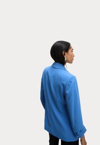 Blauwe blazer met een getailleerde pasvorm, voorzien van aangeknipte revers en opgerolde mouwen. Gladde stof en gestructureerd ontwerp.