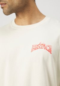 Krems krāsas kokvilnas T-krekls ar sarkano "BENFICA" logotipu kreisajā krūts daļā. Klasisks apaļais kakls, īsas piedurknes un mīksta tekstūra.