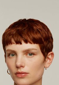 Cabello corto y texturizado de color marrón rojizo con un flequillo, con una vista en primer plano de un rostro con ojos azul-verdes y pequeños aretes de plata en forma de aro.