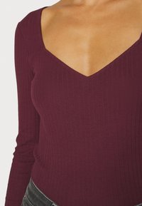 Burgunderfarbener, gerippter Langarmoberteil mit tiefem V-Ausschnitt und figurnahem Schnitt. Texturierter Stoff mit glatter Oberfläche.