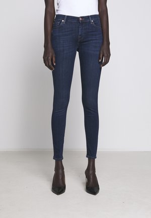 Osoba ubrana w ciemnoniebieskie obcisłe dżinsy, białą bluzkę i czarne czółenka na wysokim obcasie z ostrym noskiem, stojąca na jednolitym jasnym tle.