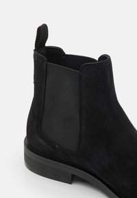 Schwarze Wildleder-Stiefelette mit elastischen Seiteneinsätzen, einer flachen schwarzen Sohle und einer Zuglasche auf der Rückseite für ein einfaches An- und Ausziehen. Strukturierte Oberfläche.