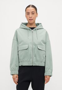Veste à capuche vert clair avec fermeture éclair, poignets et ourlet élastiqués, dotée de deux poches avant et d'une texture lisse et brillante.