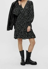 Robe noire à fleurs avec des manches longues, décolleté en V et jupe en volants. Associée à des bottes noires jusqu'aux genoux avec une semelle épaisse.