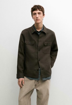 Jonge man die een donkerbruine jas met knopen draagt over een blauw shirt en beige broek, staand tegen een eenvoudige lichte muur.