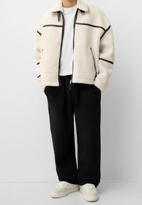 Fuzzy krämfärgad zip-up jacka med svarta accenter, matchad med svarta byxor och ljusa sneakers. Jackan har krage och sidofickor.