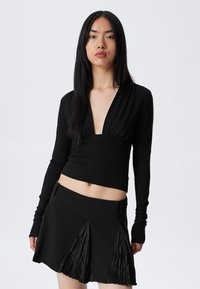 Young woman with long black hair wearing a black deep V-neck long-sleeve top and a black skirt with pleated panels.