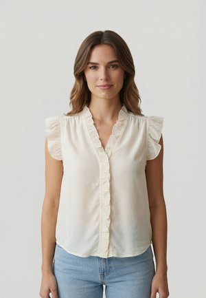 Femme aux cheveux bruns ondulés portant un chemisier sans manches couleur crème avec des bords à volants et un jean bleu clair, debout devant un fond clair uni.