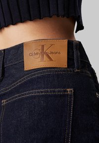 Jeans de denim oscuro con un parche de cuero marrón que presenta el logo "Calvin Klein Jeans". Costuras en contraste y un diseño ajustado.
