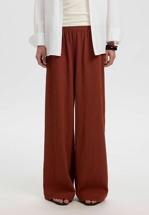 Pantalon large couleur rouille porté avec une chemise blanche boutonnée et des sandales noires à bout ouvert, mains détendues le long du corps.