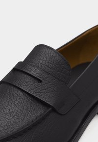 Mocassin en cuir noir texturé avec une bride à monnaie et une doublure intérieure beige, vue en gros plan sur un fond blanc.