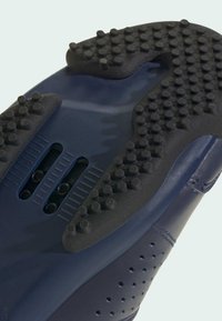 Lähikuva tummansinisestä kengänpohjasta, jossa on tekstuurilla varustettu kumikuviointi ja ilmanvaihtoaukot vaalealla taustalla.