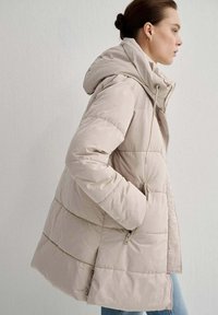 Giacca puffer beige con cappuccio, caratterizzata da un design trapuntato, cerniere laterali e dettagli con cordoncino. Texture liscia e taglio di media lunghezza.