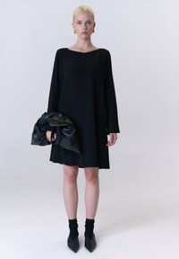 Schwarzes, langärmeliges Kleid mit quadratischem Ausschnitt, kombiniert mit einer schwarzen, geknoteten Handtasche und spitzen schwarzen Schuhen. Glattes Material, minimalistisches Design.