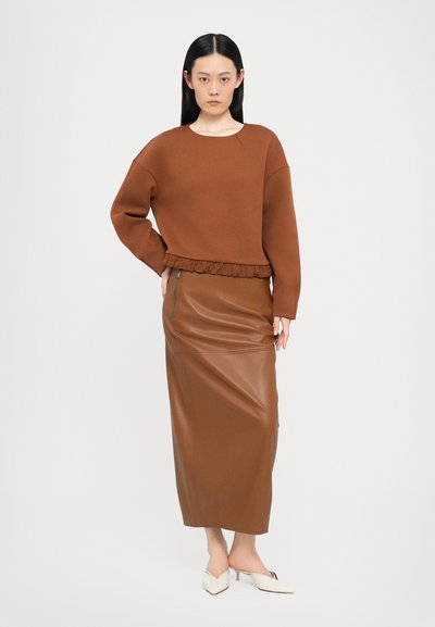 Rjava skrajšana pulover z nabranim robom, v kombinaciji z rjavim maksi krilom iz umetnega usnja, ki ima stranski zadrgo. Bele špičaste mule.