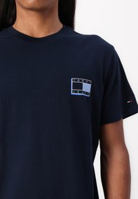 T-shirt en coton bleu marine à manches courtes, avec un patch logo à l'avant en bleu clair portant l'inscription "TOMMY JEANS." Design simple, texture épurée.