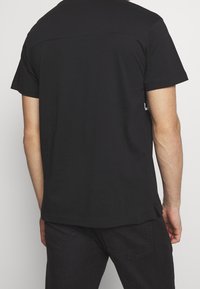 Mies, jolla on yksinkertainen musta lyhythihainen t-paita ja mustat housut, näkyy takaa yksinkertaista vaaleaa taustaa vasten.