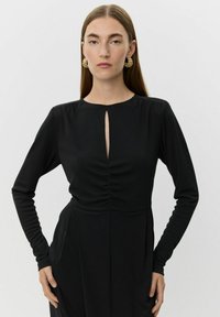 Kvinde med langt, glat brunt hår iført guldringe i ørene og en sort kjole med lange ærmer, frontskæring ved brystet og rynket talje.