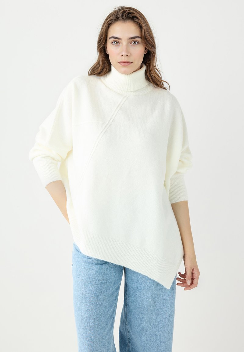 Pull blanc oversize avec col roulé et ourlet asymétrique. Texture tricotée avec poignets côtelés. Porté sur un jean large bleu clair.