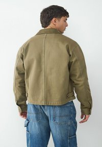 Olive green, cropped jacket made of sturdy fabric; features a collared neck, button cuffs, and seam detailing. Paired with loose blue jeans.