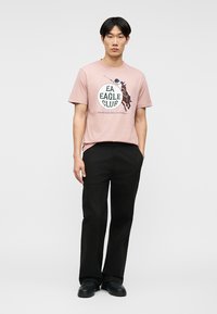 Άνδρας όρθιος με τα χέρια στις τσέπες, φοράει ροζ μπλουζάκι με σχέδιο παίκτη πόλο και μαύρο παντελόνι, σε απλό λευκό φόντο.