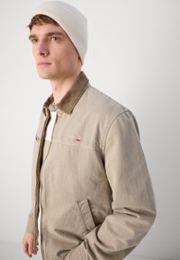 Chaqueta bomber beige con cuello marrón, presenta un cierre delantero, un bolsillo y un logo rojo. Gorro blanco llevado en la cabeza.