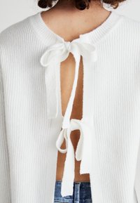 White knitted cardigan with a ribbed texture, featuring a back opening tied with white fabric ribbons, open to the mid-back.