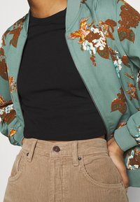 Chaqueta bomber verde con bordado floral, top corto negro y pantalones de pana beige. Presenta puños elásticos y cierre de cremallera.