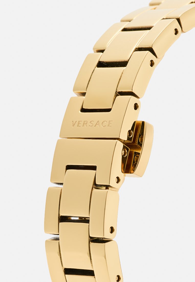 Montre-bracelet en métal doré avec un design massif, comportant des maillons rectangulaires et un fermoir gravé "Versace". Fini lisse et poli.