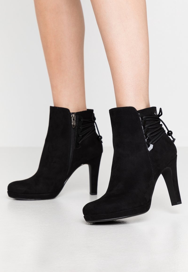Bottines en daim noir avec fermetures éclair latérales et détails lacés à l'arrière, portées sur jambes nues avec talons aiguilles hauts.