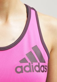 Różowy sportowy top na ramiączkach z ciemnoszarym obszyciem i dużym ciemnoszarym logo Adidas. Gładki materiał z designem typu racerback.