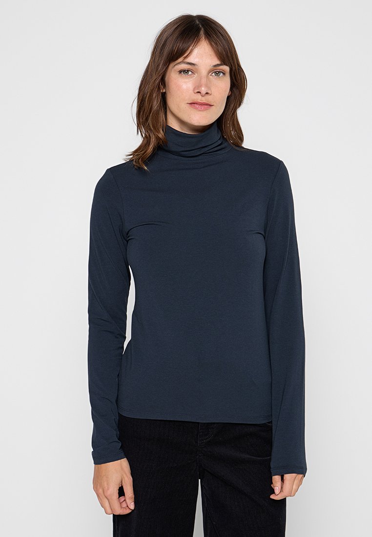 Vrouw met schouderlang bruin haar, gekleed in een donkerblauwe turtleneck met lange mouwen en zwarte broek, staand tegen een witte achtergrond.