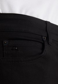 Los jeans de mezclilla negra presentan un bolsillo delantero con un logo sutil, costuras en contraste y una textura suave. La pretina es parcialmente visible.