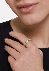 Goldener Ring mit mehreren rechteckigen klaren Steinen, die in einem gebogenen Band eingesetzt sind, getragen an einer Hand mit glatter Textur vor einem dunklen Hintergrund.