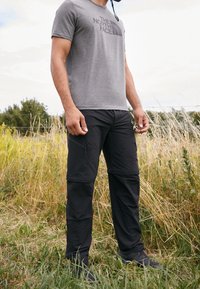 Camiseta gris de manga corta con logo, pantalones cargo negros desmontables, entorno al aire libre con hierba alta y arbustos, diseño minimalista y funcional.