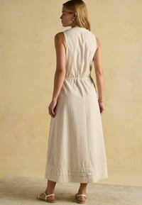 Prúžkované maxi šaty bez rukávov v krémovej a béžovej farbe; majú elastický pás a skladaný lem. Sandále dopĺňajú outfit.